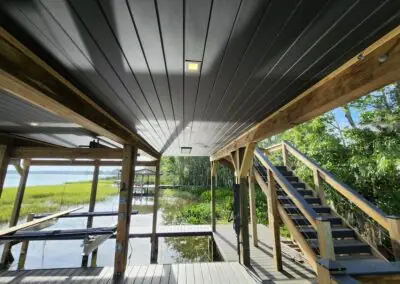Under a covered boathouse: dark ceiling, wooden beams and stairs to the upper deck, lake beyond